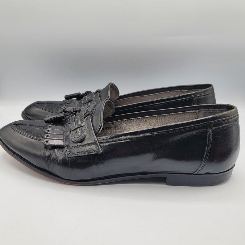 May include: Pair of black leather loafers with a glossy finish. The shoes feature a decorative tassel and fringe detail on the vamp. The soles are a dark brown color.