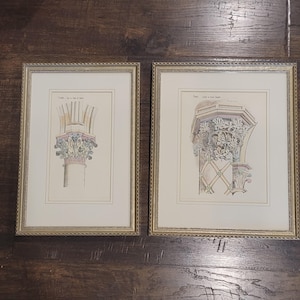 May include: Two framed architectural prints. Each print features a detailed, colourful illustration of a column capital. The frames are gold with a textured border. The prints are on a light cream-coloured background.