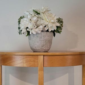 May include: A decorative arrangement of white flowers in a textured, gray ceramic pot. The flowers are a mix of different shapes and sizes, with green accents. The pot sits on a light brown, half-circle wooden table.