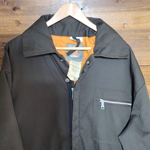 May include: A brown work jacket with a collar, zipper, and a chest pocket with a silver zipper. The jacket has an orange quilted lining and a tag that reads "HAMMER". The jacket is hanging on a wooden hanger.