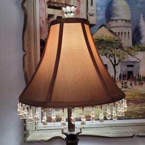 May include: A decorative lamp with a brown lampshade and a dark brown trim. The lampshade is decorated with clear crystal beads. The lamp has a floral design at the top and is set against a painting.