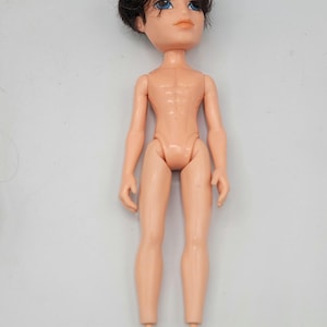 Könnte beinhalten: Eine Bratz-Puppe mit gebräuntem Teint, blauen Augen und kurzen, dunkelbraunen Haaren. Die Puppe ist nackt und hat einen muskulösen Körperbau. Die Puppe steht aufrecht vor einem weißen Hintergrund.