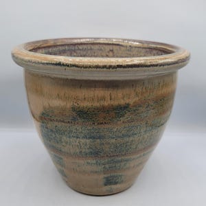 Puede incluir: Una maceta de cerámica con un borde ancho y redondeado y un cuerpo cónico. El exterior presenta una mezcla de tonos terrosos, incluyendo beige, marrón y verde azulado, con un acabado texturizado y esmaltado. Para uso en interiores o exteriores.