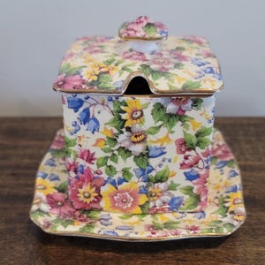 May include: A floral-patterned ceramic sugar bowl with a lid and a matching saucer. The square bowl has a small opening for a spoon. The design features pink, yellow, and blue flowers on a cream background, with gold trim.
