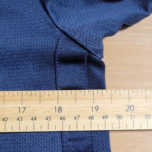 Puede incluir: Primer plano de una camisa azul marino con tejido de malla. Una regla de madera se coloca debajo de la camisa, mostrando medidas de 42 a 52 pulgadas.