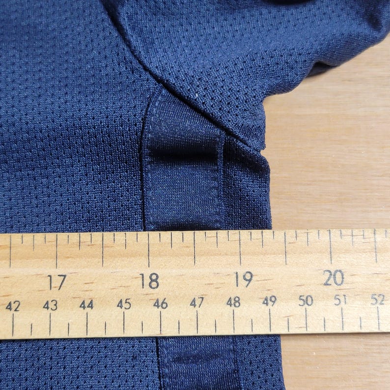 Puede incluir: Primer plano de una camisa azul marino con tejido de malla. Una regla de madera se coloca debajo de la camisa, mostrando medidas de 42 a 52 pulgadas.