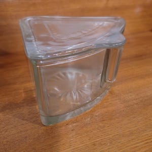 May include: A clear, triangular glass block with a curved top and a decorative, starburst-like pattern on the front. The block is designed with a handle on one side and sits on a wooden surface.
