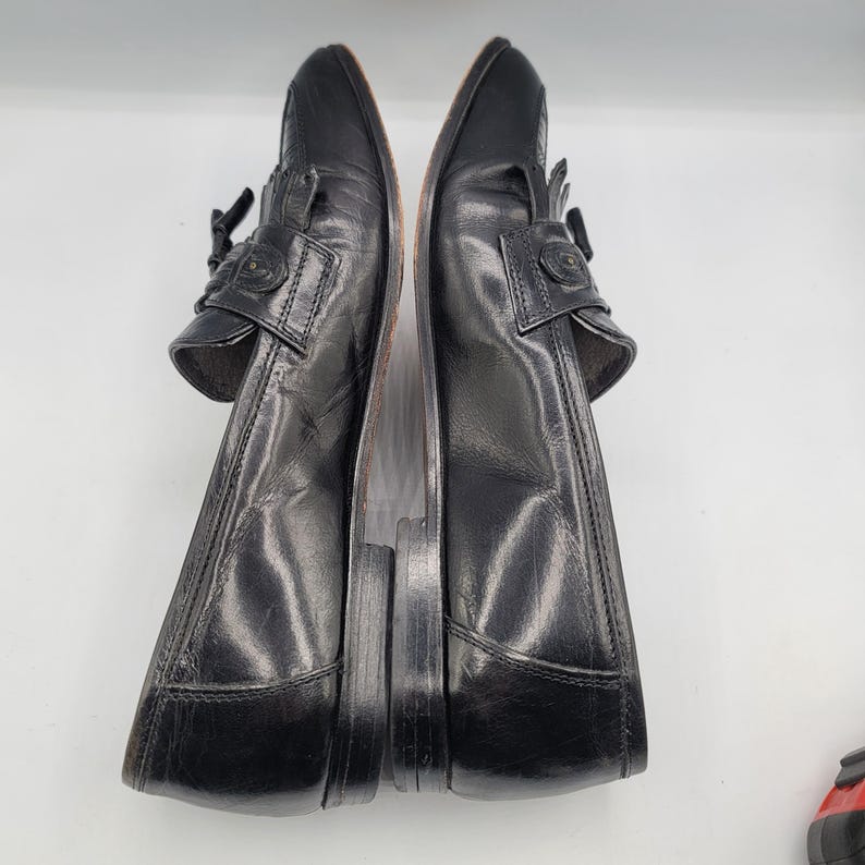 May include: Pair of black leather loafers with decorative accents. The shoes have a classic design with a rounded toe and a low heel. The leather appears to be slightly worn, adding to the vintage aesthetic.