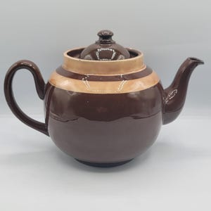May include: A brown ceramic teapot with a matching lid. The teapot has a rounded body with a tan stripe around the middle. It features a curved handle and a spout for pouring. The teapot is a classic design.