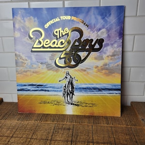 Könnte beinhalten: Ein gold-schwarzes Programmcover für die 50. Jubiläumstour der Beach Boys. Das Cover zeigt einen Sonnenuntergang über dem Meer mit einer Silhouette einer Person, die auf einem Pferd reitet.