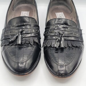 May include: Pair of black leather loafers with decorative tassels and fringe. The shoes have a textured pattern on the toe and a leather sole. The shoes are in good condition and appear to be well-maintained.