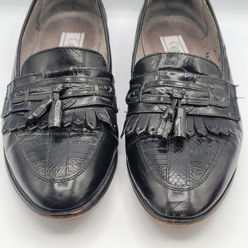 May include: Pair of black leather loafers with decorative tassels and fringe. The shoes have a textured pattern on the toe and a leather sole. The shoes are in good condition and appear to be well-maintained.