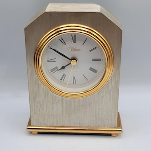 Pode incluir: Um relógio de mesa prateado e dourado com numerais romanos e a palavra "Richeau" no mostrador. O relógio é de quartzo.