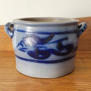 May include: A cylindrical stoneware crock featuring a light gray glaze with cobalt blue hand-painted floral motifs and horizontal bands. The vessel includes two side handles and a wide, open top rim, showcasing a salt-glazed texture throughout.