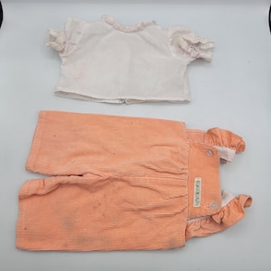 Może przedstawiać: Zestaw ubrań dla niemowląt w stylu vintage, składający się z białej bluzki z koronkowym wykończeniem i brzoskwiniowych sztruksowych ogrodniczek. Bluzka ma krótkie rękawy i falbankowy dekolt. Ogrodniczki mają zapięcie na guziki i falbankowy dół. Zestaw jest prawdopodobnie dla dziecka lub malucha.