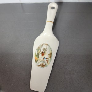 May include: White ceramic cake server with a gold trim. The server features a winter scene with a horse-drawn carriage and the text "Christmas Greetings".