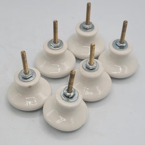 May include: A set of six white ceramic cabinet knobs, each featuring a smooth, rounded top and a central metallic threaded bolt with a silver-coloured nut and washer assembly attached to the top for secure installation on furniture or cabinetry.