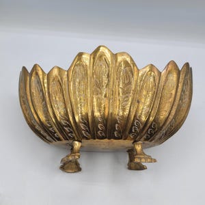 Può includere: Una ciotola in ottone color oro con bordo smerlato e un motivo floreale inciso su ogni sezione a forma di petalo. La ciotola è sostenuta da tre piedi decorati a forma di artiglio, che forniscono una base rialzata al corpo arrotondato.