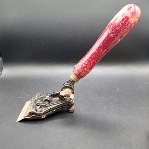 May include: A vintage paint scraper featuring a weathered red wooden handle attached to a black metal head. The tool includes an adjustable hinge mechanism with a wing nut, a flat metal blade, and a small support foot for stability during use.