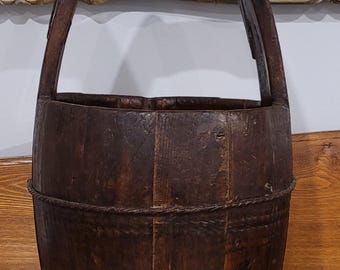 Puede incluir: Un cubo de madera marrón oscuro con un asa. El cubo está hecho de tablones de madera verticales sujetos con cuerda. El asa es arqueada y está unida a la parte superior del cubo. El cubo se encuentra sobre una superficie de madera.