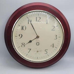 May include: A round wall clock with a deep red frame and a cream-coloured face. The clock features black Arabic numerals, a silver-toned inner bezel, and three black hands. A small sub-dial for seconds is positioned at the bottom with the text Quartz.
