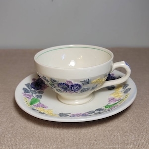 Puede incluir: Juego de taza y platillo de té color crema con un diseño floral en morado, azul y amarillo. La taza tiene un borde verde y un acento azul en el asa. El platillo refleja el patrón floral.