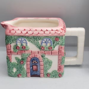 May include: A ceramic creamer shaped like a cottage with a pink scalloped roof, white walls, and a blue arched door. The exterior features green climbing vines with small red flowers, two windows with blue frames, and a white rectangular handle.