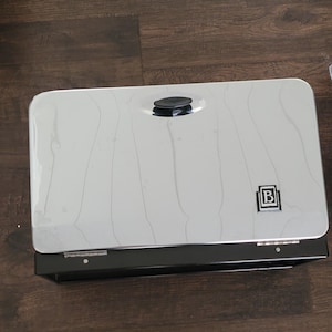 Peut inclure: Une boîte à pain rectangulaire en acier inoxydable et en métal noir. Le couvercle est blanc avec un motif subtil marbré et une poignée noire. La boîte a une base noire et un logo dans le coin inférieur droit.