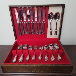 Puede incluir: Un juego de cubiertos vintage en una caja de madera con interior de terciopelo rojo. El juego incluye cuchillos, tenedores, cucharas y cucharas de servir. La cubertería tiene mangos ornamentados y una superficie brillante y reflectante.