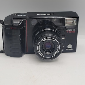 May include: A black Minolta AF-Tele 35mm film camera featuring a dual-lens system with 38mm and 60mm focal lengths. The front of the camera displays the Minolta logo, an integrated flash, a shutter button, and the text Auto Focus Tele.