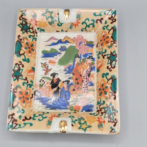 Op de afbeelding: Een vierkante asbak van keramiek met een gouden rand en een kleurrijk bloemenontwerp. Het midden van de asbak toont een scène met vier vrouwen in traditionele Japanse kleding die in een landschap met bergen en water staan.