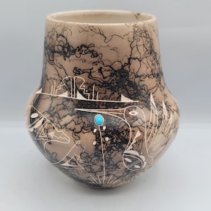 May include: A tan ceramic vase featuring black, lightning-like patterns and white etched designs. A small turquoise stone is set into the side. The vase has a rounded body and a slightly flared rim.