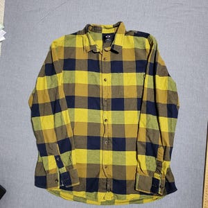 May include: A long-sleeved, button-down shirt in a plaid pattern of yellow, navy blue, and brown. The shirt has a collar, buttoned cuffs, and a small logo on the chest. The shirt is made of a soft, woven fabric.
