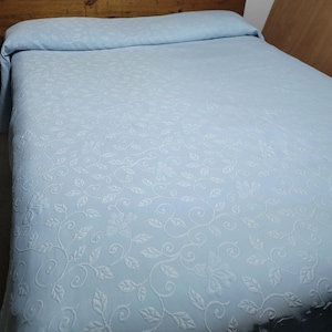 May include: A light blue bedspread featuring a tonal, raised floral and vine pattern across the entire surface. The fabric has a textured appearance with swirling stems and leaf motifs that repeat consistently over the rectangular bedding.