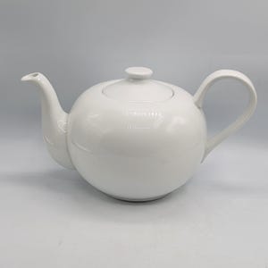 May include: A white ceramic teapot with a rounded body, a curved spout, a matching lid with a small knob handle, and a C-shaped handle. The teapot features a smooth, glossy finish and a clean, simple design with no additional patterns or colours.