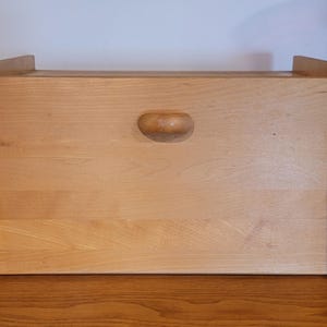 May include: A rectangular light brown wooden bread bin featuring a smooth finish, a drop-down front panel, and a small, curved wooden handle centred on the front. The structure is framed by two vertical side panels with rounded top edges.