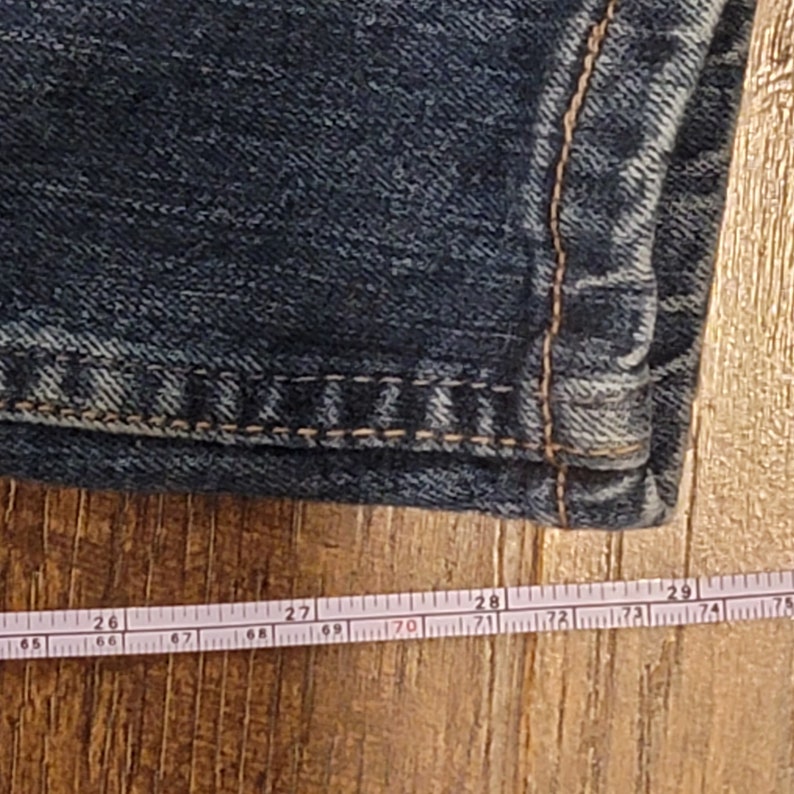 Puede incluir: Primer plano del borde inferior de un par de pantalones vaqueros de color azul oscuro. Una cinta m&eacute;trica blanca est&aacute; colocada sobre una superficie de madera, mostrando medidas de 66 a 74 cent&iacute;metros.