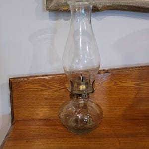 May include: A clear glass oil lamp featuring a bulbous base with a textured hobnail pattern, a brass burner assembly, and a tall, slender, transparent glass chimney. The lamp is constructed with a wide, rounded bottom section and a narrow neck.