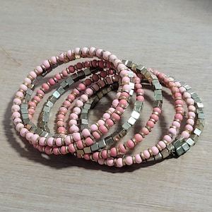 Puede incluir: Conjunto de seis pulseras elásticas. Cada pulsera presenta una combinación de pequeñas cuentas redondas de color rosa claro, pequeñas cuentas doradas y cuentas cuadradas plateadas. Las pulseras están dispuestas en un patrón circular sobre una superficie de madera clara.