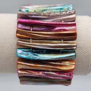 Puede incluir: Un brazalete de concha iridiscente multicolor con un diseño apilado. El brazalete está hecho de muchas piezas individuales de concha que están apiladas una encima de la otra. Las piezas de concha son de diferentes colores, incluyendo rosa, morado, azul, verde y marrón. El brazalete es una pieza de joyería hermosa y única.