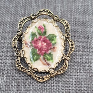 Könnte beinhalten: Vintage goldfarbene Brosche mit einem ovalen, kreuzgestickten Blumendesign. Die Stickerei zeigt eine rosa Rose mit grünen Blättern und Knospen. Die Brosche hat einen verzierten Rahmen im Filigranstil.