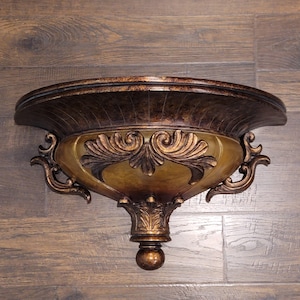 May include: An ornate wall shelf in bronze and gold tones. The shelf has a curved top and a decorative, vase-like body with scrollwork and a rounded base. Designed to be mounted on a wall.