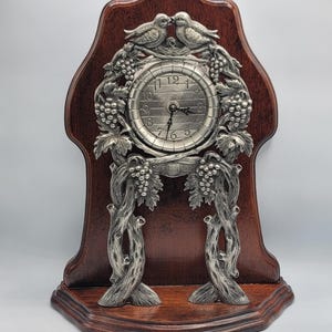 May include: A mantel clock featuring a dark brown wooden arched backboard and a circular silver-toned metal face. The clock is framed by intricate silver-toned metal relief carvings of grapevines, clusters of grapes, and two birds perched at the top.