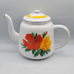 Könnte beinhalten: Weiße Emaille-Teekanne mit Deckel und Griff. Die Teekanne ist mit roten und gelben Blumenmustern und grünen Blättern verziert. Der Deckel hat einen gelben Streifen. Ausguss und Griff sind weiß.