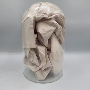 May include: A clear glass cloche with a rounded top, filled with beige-coloured crumpled paper. The cloche sits on a clear, circular base. The background is a solid light grey.