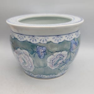 May include: A round ceramic planter featuring a muted sage green background with a repeating pattern of light purple and white flowers and butterflies. The rim and base are decorated with a blue and white scalloped border design.