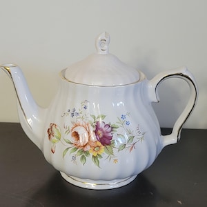 May include: A white porcelain teapot with a floral pattern of pink, purple, and yellow roses. The teapot has a curved spout, a loop handle, and a lid with a small handle. Gold trim edges the teapot.