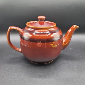 May include: A ceramic teapot in shades of brown, featuring a lid and handle. The main body is a deep reddish-brown, with a darker band around the middle. The spout and handle are a lighter brown. Designed for brewing tea, it's a classic kitchenware item.