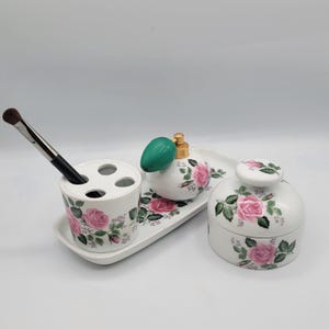 May include: White porcelain bathroom set with a rose floral design. The set includes a toothbrush holder, a tray, a small bottle with a green lid, and a round jar with a lid.