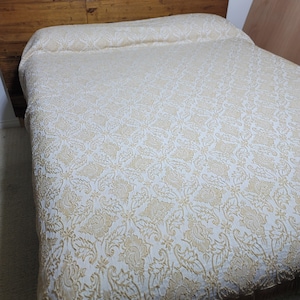 May include: A bedspread featuring a cream and light yellow damask pattern with intricate floral and leaf motifs. The fabric has a textured, woven appearance that covers the entire surface of the bed, extending from the headboard to the foot.